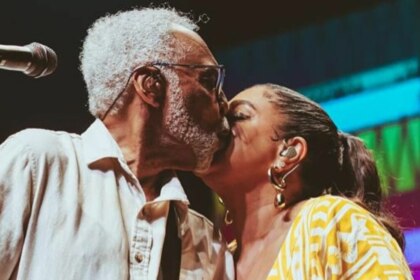 Gilberto Gil, com barba e cabelos grisalhos, beija a testa de sua filha, Preta Gil. Ambos estão em um palco. Gilberto usa uma camisa branca e óculos, enquanto Preta, com os cabelos presos, usa brincos dourados. Um microfone está visível à esquerda.
