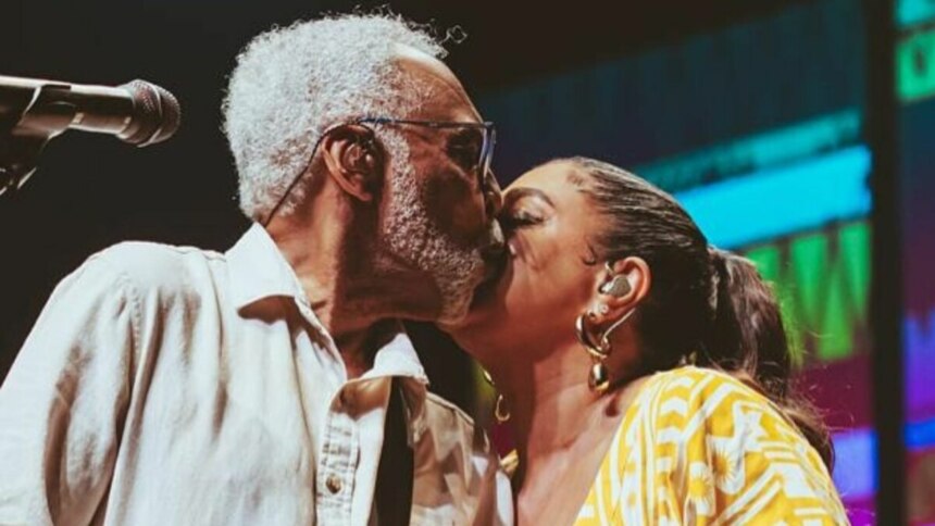 Gilberto Gil, com barba e cabelos grisalhos, beija a testa de sua filha, Preta Gil. Ambos estão em um palco. Gilberto usa uma camisa branca e óculos, enquanto Preta, com os cabelos presos, usa brincos dourados. Um microfone está visível à esquerda.