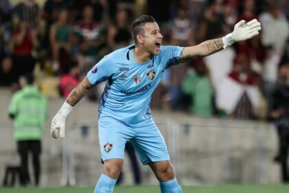 A imagem mostra o goleiro Fábio, do Fluminense, durante uma partida de futebol. Ele usa o uniforme de goleiro na cor azul-claro, com o logo da Umbro e patrocínio da Superbet. Ele tem os braços abertos e as luvas brancas, gritando com uma expressão séria. É possível ver as tatuagens em seus braços. O fundo mostra a arquibancada com torcedores.