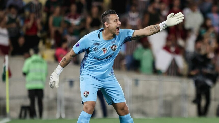 A imagem mostra o goleiro Fábio, do Fluminense, durante uma partida de futebol. Ele usa o uniforme de goleiro na cor azul-claro, com o logo da Umbro e patrocínio da Superbet. Ele tem os braços abertos e as luvas brancas, gritando com uma expressão séria. É possível ver as tatuagens em seus braços. O fundo mostra a arquibancada com torcedores.