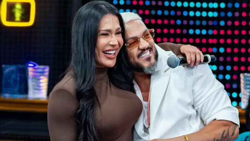 A imagem mostra a atriz e influenciadora Gracyanne Barbosa e o cantor Belo juntos, abraçados, em um estúdio de televisão. Gracyanne sorri, com a cabeça apoiada no ombro de Belo. Ela usa um vestido marrom. Belo também sorri, com a mão direita no ombro de Gracyanne e a esquerda segurando um microfone. Ele usa um casaco branco e óculos de sol com lentes laranjas. Ao fundo, um painel com luzes coloridas e um neon amarelo.