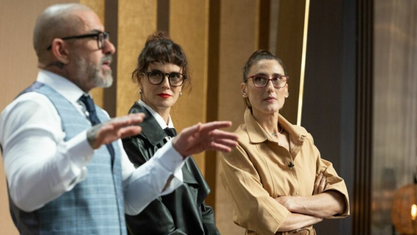 Os três jurados do programa MasterChef Brasil em pé na cozinha do estúdio. Henrique Fogaça, à esquerda, gesticula com as mãos enquanto fala. Helena Rizzo e Paola Carosella, no centro e à direita, respectivamente, o observam com expressões sérias.