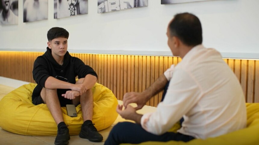 A imagem mostra uma cena do programa "Globo Repórter". À esquerda, um garoto de cabelos escuros está sentado em um pufe amarelo, usando um moletom preto e bermuda preta. Ele tem uma expressão séria e está com as mãos entrelaçadas. À direita, o repórter Jean Raupp está de costas para a câmera, sentado em outro pufe amarelo, com uma camisa social clara. O fundo mostra uma parede com quadros e revestimento de madeira.