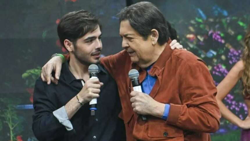 Os apresentadores João Silva e Faustão, pai e filho, em pé no palco de um programa de televisão, seguram microfones e sorriem um para o outro. Faustão, de jaqueta marrom, tem o braço sobre o ombro de João, que veste uma camisa escura. O fundo do palco está desfocado com luzes coloridas.