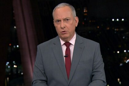 O jornalista José Roberto Burnier, usando um terno cinza, camisa rosa clara e gravata vermelha, apresenta o jornal. Ele olha seriamente para a câmera, com um fundo desfocado que mostra o cenário do telejornal "SP2" e as luzes de uma cidade à noite.