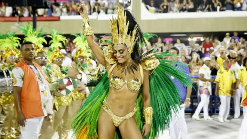 A atriz Juliana Paes, vestida com uma fantasia exuberante de rainha de bateria, nas cores dourado e verde, dança com os braços erguidos durante um desfile de carnaval. Ela sorri com os olhos fechados. Ao fundo, é possível ver outras pessoas fantasiadas e a plateia do sambódromo.