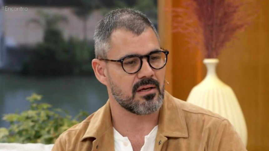 O ator Juliano Cazarré em um close-up, sentado no sofá do programa 'Encontro'. Ele usa óculos de armação escura, uma jaqueta marrom clara e uma camisa branca. Ele tem barba e bigode grisalhos e parece estar falando seriamente. Ao fundo, o cenário do estúdio é visível.