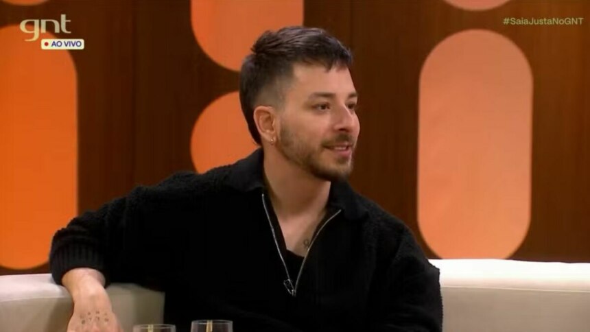 O cantor e apresentador Junior Lima sentado em um sofá no estúdio do programa "Saia Justa". Ele usa um suéter preto de zíper, sorri discretamente e olha para o lado. O logo da emissora GNT está visível no canto superior da tela.