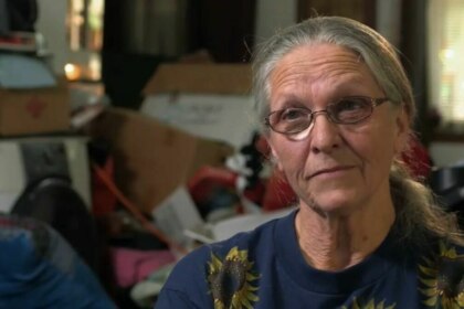 Mulher de cabelos grisalhos e óculos, com uma camiseta de girassol, olha para a câmera com um sorriso sutil. O fundo está desfocado e mostra um ambiente bagunçado, com caixas e objetos empilhados, em trecho do programa Acumuladores