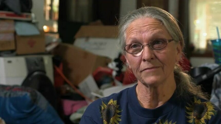 Mulher de cabelos grisalhos e óculos, com uma camiseta de girassol, olha para a câmera com um sorriso sutil. O fundo está desfocado e mostra um ambiente bagunçado, com caixas e objetos empilhados, em trecho do programa Acumuladores