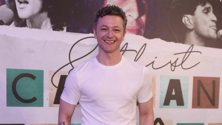O músico Lucas Lima, em um close-up de meio-corpo, sorri para a câmera. Ele veste uma camiseta branca lisa. O fundo da foto é um banner com letras e imagens desfocadas.