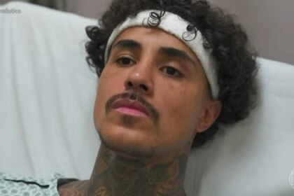 MC Livinho, homem com cabelos cacheados escuros e uma faixa branca na cabeça, deitado em uma cama de hospital, vestindo um avental hospitalar estampado. Ele tem barba e bigode finos, várias tatuagens visíveis no pescoço e olha para a frente com uma expressão séria. No canto superior esquerdo, há a hashtag "#Fantástico".