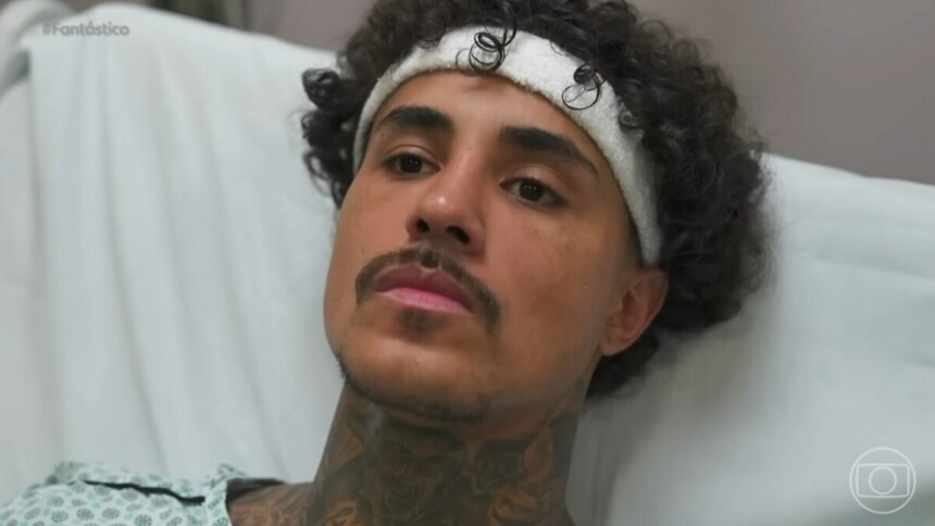 MC Livinho, homem com cabelos cacheados escuros e uma faixa branca na cabeça, deitado em uma cama de hospital, vestindo um avental hospitalar estampado. Ele tem barba e bigode finos, várias tatuagens visíveis no pescoço e olha para a frente com uma expressão séria. No canto superior esquerdo, há a hashtag "#Fantástico".
