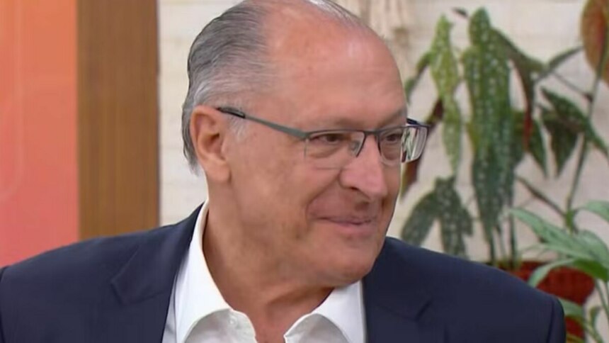 Um close-up de Geraldo Alckmin, um homem idoso de pele clara, com pouco cabelo grisalho penteado para trás. Ele usa óculos de armação fina e um terno azul marinho com uma camisa branca aberta no colarinho, em participação no Mais Você. Ele está olhando para a direita da imagem com uma expressão facial levemente sorridente e um fundo desfocado de plantas e objetos decorativos.