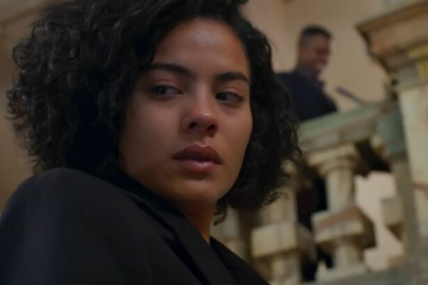 A imagem é um close da atriz Bella Campos, interpretando a personagem Maria de Fátima na novela "Vale Tudo". Ela tem cabelos curtos e cacheados, e sua expressão facial é séria, com o olhar desviado para o lado. Ela está usando uma jaqueta preta. Ao fundo, desfocado, é possível ver um homem em um ambiente interno, com colunas de pedra.