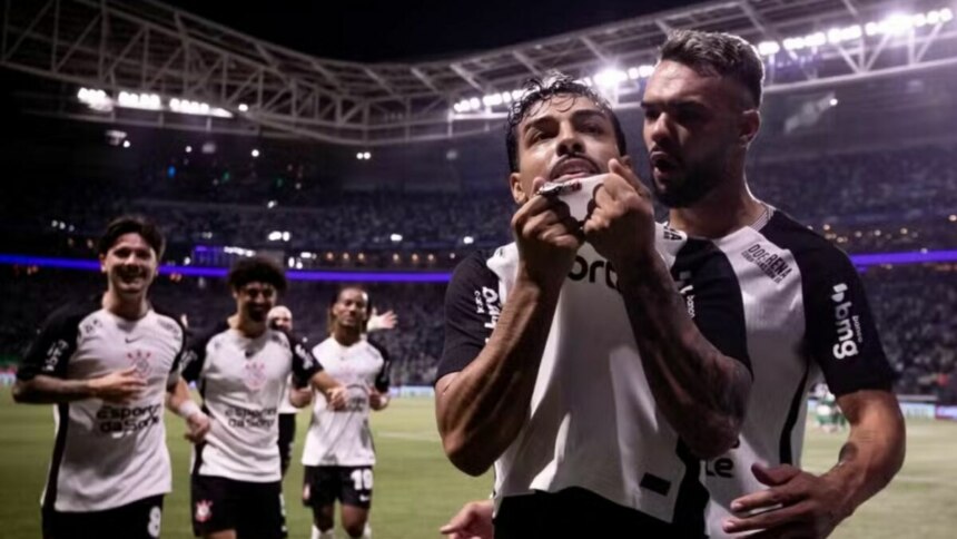 O jogador de futebol Matheus Bidu, do Corinthians, celebra um gol em um estádio lotado contra o Palmeiras. Ele puxa a camisa sobre o rosto e olha para cima, enquanto um colega de time o abraça. Outros jogadores do Corinthians correm para perto para comemorar, e as luzes do estádio iluminam a cena noturna.
