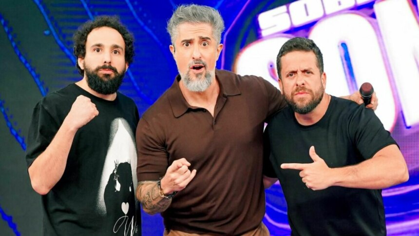 Os humoristas Maurício Meirelles e Murilo Couto, e o apresentador Marcos Mion, posam juntos no palco do programa Caldeirão. Mion, ao centro, tem uma expressão de surpresa.