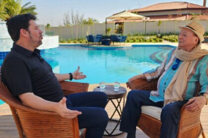 A imagem mostra uma cena do programa "Domingo Espetacular". O repórter Michael Keller e o apresentador Raul Gil estão sentados em cadeiras de vime, de frente um para o outro, em uma área externa com uma piscina. Michael Keller, à esquerda, veste uma camisa polo preta e calça escura, e gesticula com as mãos enquanto fala. Raul Gil, à direita, usa um chapéu de palha, uma camisa listrada e um cachecol. Entre eles, há uma pequena mesa com dois copos de água. O fundo é uma casa e uma área de lazer.