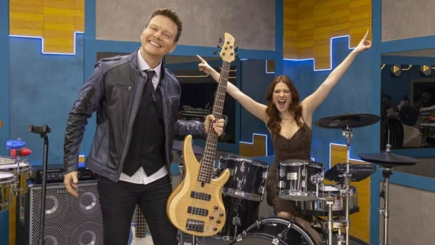 A imagem mostra Michel Teló e Ana Clara no cenário musical do programa Estrela da Casa. Michel Teló, à esquerda, sorri e segura um baixo elétrico. Ele usa uma jaqueta de couro azul-marinho, um colete preto e uma gravata. Ana Clara, à direita, está sentada atrás de uma bateria, com os braços erguidos em comemoração, e usa um vestido marrom. Ao fundo, o cenário é composto por formas geométricas em tons de azul e amarelo, com luzes de neon.