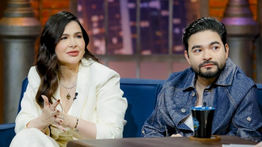 Mila Braga e Yudi Tamashiro sentados em um sofá azul no estúdio do programa 'The Noite'. Mila, de cabelo escuro, usa um blazer branco e está com as mãos unidas. Yudi, com barba e jaqueta jeans, também está sentado ao lado dela. Ambos parecem estar em uma entrevista.