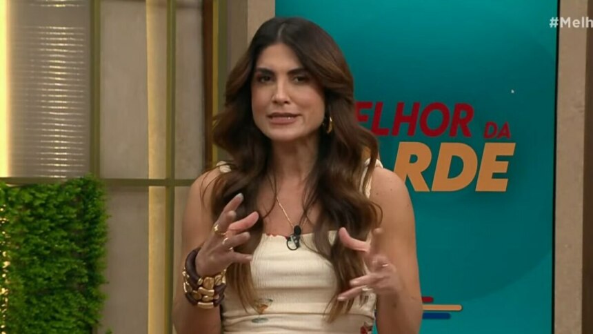 A imagem é um close da apresentadora Pâmela Lucciola, em uma cena do programa "Melhor da Tarde". Ela está em pé, gesticulando com as mãos enquanto fala. Pâmela usa uma blusa de alças bege com detalhes e braceletes e anéis dourados. Ao fundo, um painel com a inscrição "MELHOR DA TARDE" e o logo do programa.