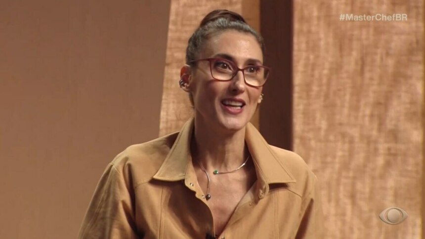 A chef e jurada Paola Carosella em um close-up, sorrindo e falando em uma cena do programa "MasterChef Brasil". Ela usa óculos de armação vermelha, uma camisa bege e um colar. O fundo do estúdio é de painéis de madeira. O logo da Band e a hashtag #MasterChefBR estão visíveis na tela.
