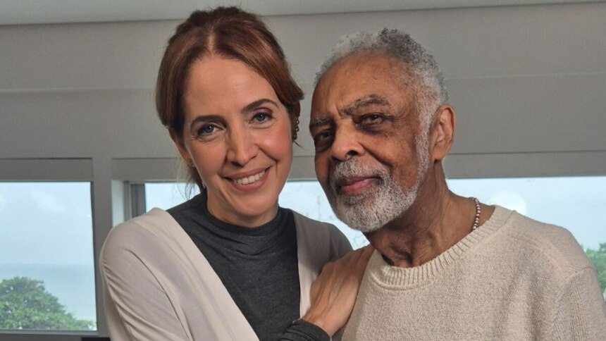 A jornalista Poliana Abritta e o cantor Gilberto Gil posam juntos em um ambiente claro. Poliana tem o braço sobre o ombro de Gilberto, e ambos sorriem levemente para a câmera. Ao fundo, grandes janelas revelam uma vista para o mar. A imagem é de uma entrevista para o programa "Fantástico".