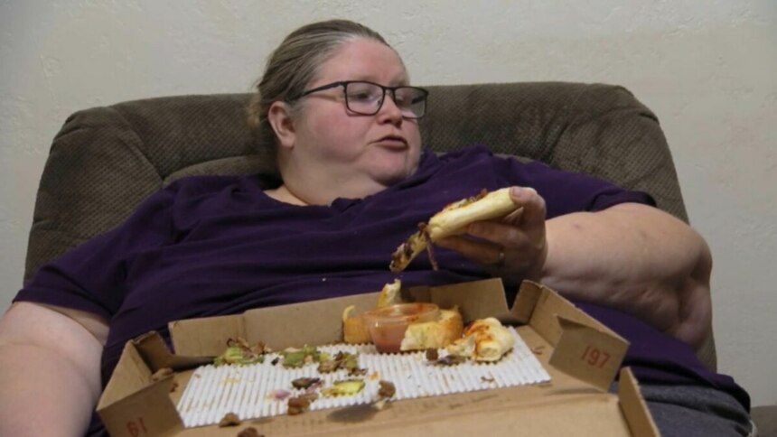 Mulher chamada Bethany, sentada em uma poltrona, segura um pedaço de pizza na mão. Ela está usando uma blusa roxa e óculos. Em seu colo, há uma caixa de pizza com restos de comida. O nome do arquivo a identifica como participante do programa 'Quilos Mortais'.