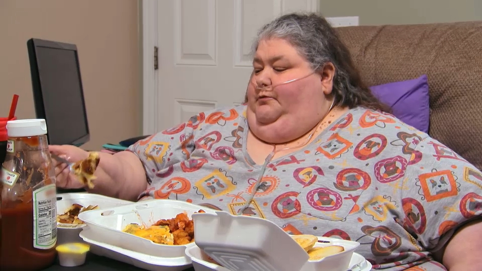 Joyce, do Quilos Mortais, sentada com oxigênio nasal e refeição à frente, durante tratamento em casa.