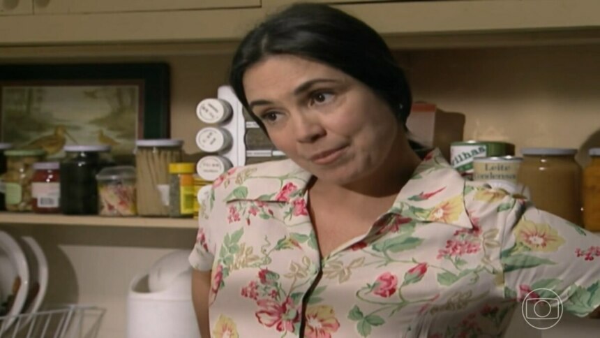 A atriz Regina Duarte, interpretando a personagem Helena, em uma cena da novela "História de Amor". Ela está em uma cozinha, usando uma blusa de estampa floral, e olha para o lado com um sorriso sutil. Ao fundo, prateleiras de madeira com utensílios e potes de temperos.