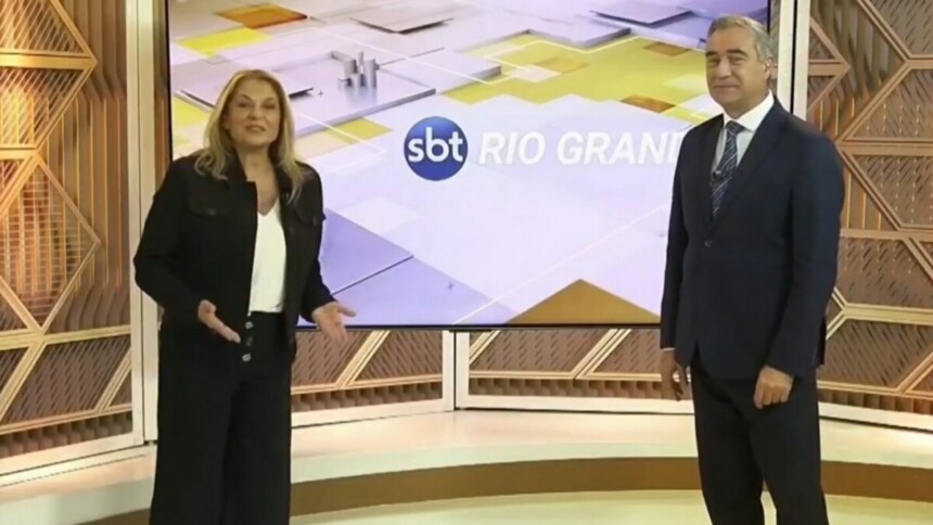 A imagem mostra a dupla de apresentadores Regina Lima e André Haar, em pé, em um estúdio de televisão. Regina, à esquerda, veste um blazer preto, calça preta e uma blusa branca. Ela gesticula com as mãos enquanto fala. André, à direita, usa um terno azul-marinho com gravata. Entre eles, há um grande painel digital exibindo o logotipo "SBT RIO GRANDE".