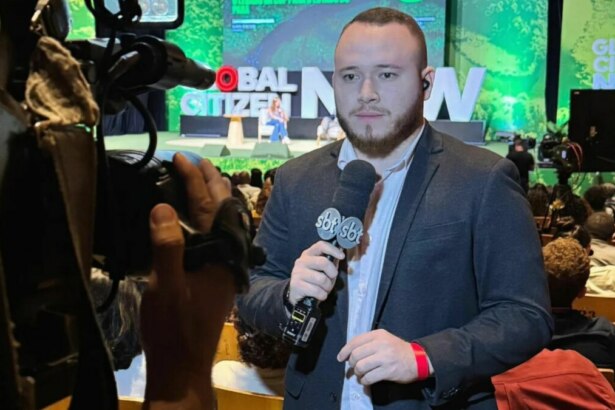 O repórter Andrey Araújo, de terno cinza, segura um microfone com o logo do SBT, enquanto olha para a câmera. Ele está em um evento com um banner 'Global Citizen' no fundo, e o nome do arquivo o associa ao SBT Pará.