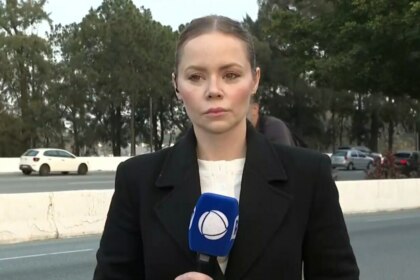 A repórter Beatriz Casadei, de casaco preto, está no acostamento de uma estrada. Ela segura um microfone com a logo da RECORD, e uma expressão séria.