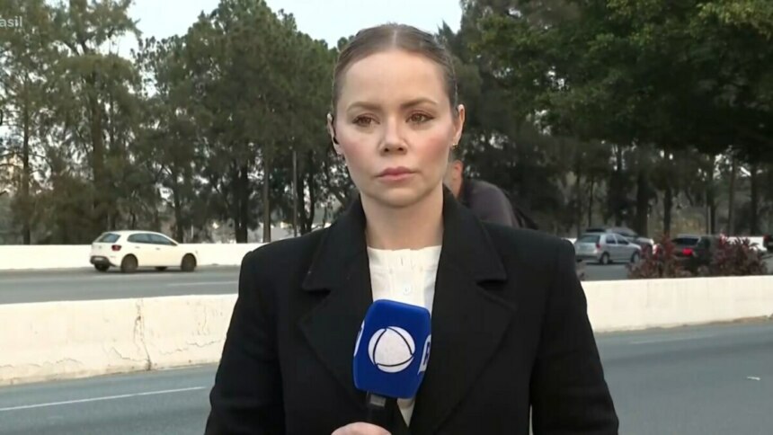 A repórter Beatriz Casadei, de casaco preto, está no acostamento de uma estrada. Ela segura um microfone com a logo da RECORD, e uma expressão séria.