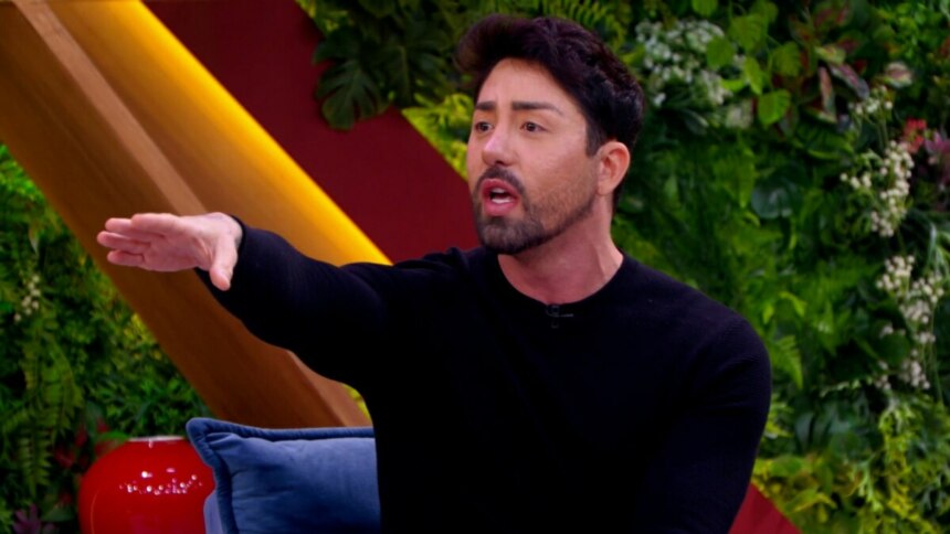 Rico Melquiades, usando um suéter preto, sentado em um sofá no programa "Casos de Família". Ele gesticula com o braço estendido e tem a boca aberta, como se estivesse falando de forma intensa. O fundo é um cenário de estúdio decorado com folhagens e painéis coloridos.