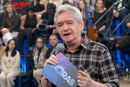A imagem é um close do apresentador Serginho Groisman, no palco do programa "Altas Horas". Ele está sorrindo, olhando para o lado, e segura um microfone com a logo do programa e um pequeno tablet. Ele veste uma camisa xadrez escura. A plateia está ao fundo, sentada em uma arquibancada com luzes azuis.