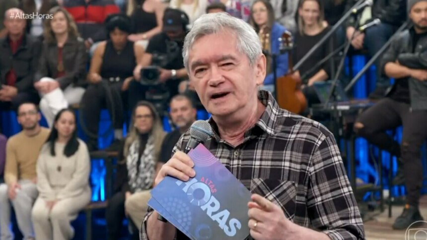 A imagem é um close do apresentador Serginho Groisman, no palco do programa "Altas Horas". Ele está sorrindo, olhando para o lado, e segura um microfone com a logo do programa e um pequeno tablet. Ele veste uma camisa xadrez escura. A plateia está ao fundo, sentada em uma arquibancada com luzes azuis.