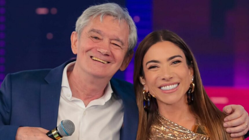 Os apresentadores Serginho Groisman e Patricia Abravanel sorriem juntos para a câmera no estúdio do "Programa Silvio Santos". Serginho, de terno azul, pisca o olho e segura um microfone, enquanto Patricia, de top dourado, sorri largamente.