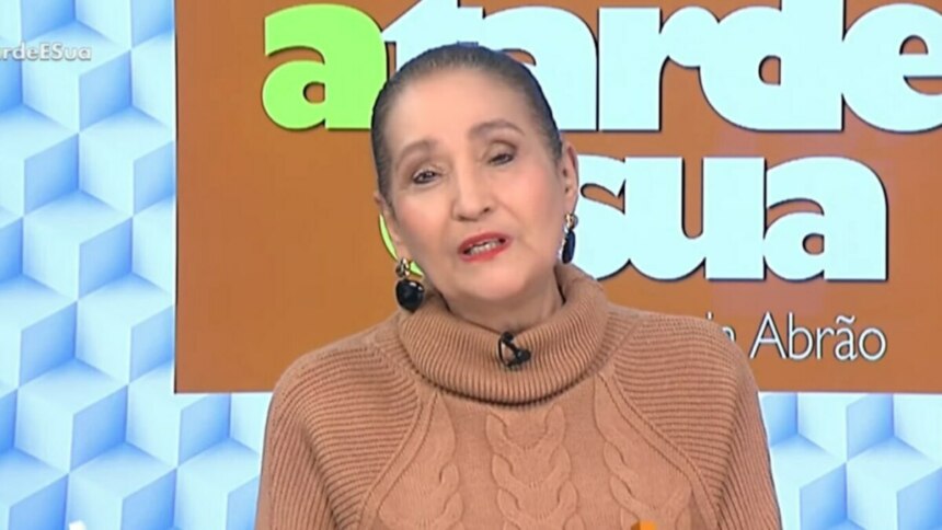A apresentadora Sonia Abrão em um estúdio de televisão, falando diretamente para a câmera. Ela usa um suéter de gola alta na cor caramelo e grandes brincos. Ao fundo, o painel do cenário exibe o nome do programa "A Tarde é Sua" e o nome dela.