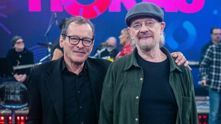 Dois integrantes da banda The Fevers posam no palco do programa "Altas Horas". À esquerda, um homem de óculos e blazer preto sorri para a câmera. Ao lado dele, um homem mais velho com barba, óculos e uma boina, sorri e tem o braço esquerdo sobre o ombro do colega. Ao fundo, parcialmente visíveis, outros músicos e equipamentos de palco em um ambiente com iluminação azulada e elementos do cenário do programa.
