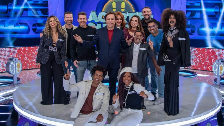 Tom Cavalcante e parte do elenco do programa "Acerte ou Caia" posam juntos em um palco iluminado. Tom, no centro, veste um terno escuro com camisa vermelha, sorrindo e apontando. Ao redor dele, diversas pessoas com figurinos variados, incluindo um homem de blazer branco agachado à frente, e uma mulher com cabelo encaracolado e roupa preta. O cenário futurista é azul com luzes vibrantes e detalhes circulares, e o público pode ser visto ao fundo.