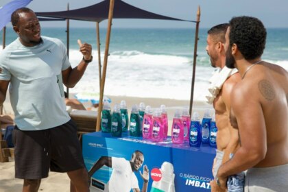A imagem mostra Usain Bolt em uma praia. Ele está de costas para a câmera, gesticulando com as mãos enquanto conversa com dois homens, um dos quais é o ator Cauã Reymond. Usain Bolt veste uma camiseta clara e shorts escuros. No centro da imagem, há um estande com garrafas de produtos OMO em várias cores e uma imagem de Usain Bolt na frente. O ator Cauã Reymond, à direita, está sem camisa e com uma toalha branca no ombro. Ao fundo, é possível ver o mar e a areia da praia, com algumas cadeiras de praia e guarda-sóis. A cena é da novela Vale Tudo na Globo.