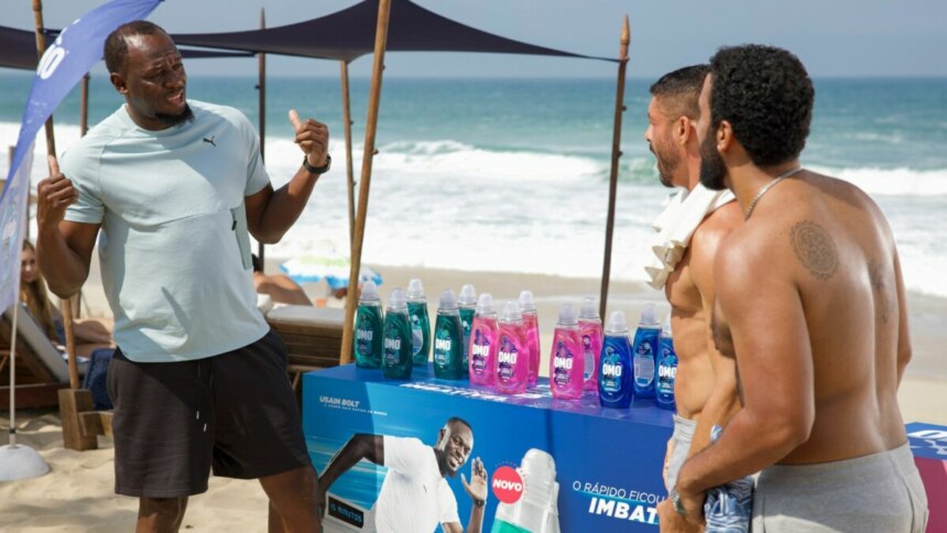 A imagem mostra Usain Bolt em uma praia. Ele está de costas para a câmera, gesticulando com as mãos enquanto conversa com dois homens, um dos quais é o ator Cauã Reymond. Usain Bolt veste uma camiseta clara e shorts escuros. No centro da imagem, há um estande com garrafas de produtos OMO em várias cores e uma imagem de Usain Bolt na frente. O ator Cauã Reymond, à direita, está sem camisa e com uma toalha branca no ombro. Ao fundo, é possível ver o mar e a areia da praia, com algumas cadeiras de praia e guarda-sóis. A cena é da novela Vale Tudo na Globo.