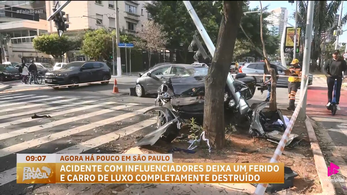 Porsche avaliado em R$ 1,5 milhão completamente destruído após colidir contra poste, em acidente com influenciadores que deixou um ferido; cena isolada por cones e fita de segurança, com presença de bombeiros e curiosos
