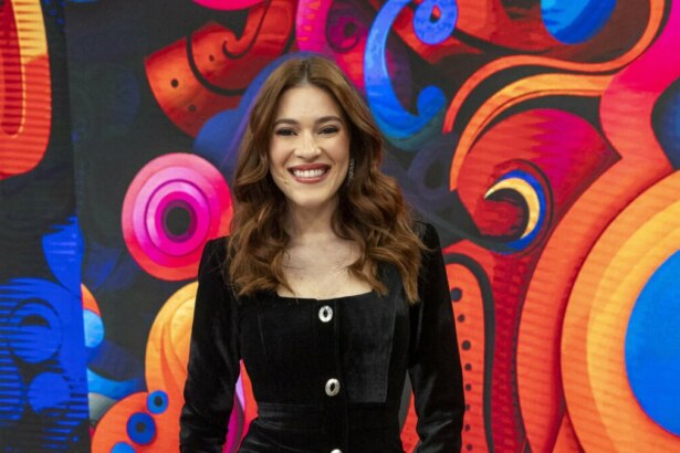 A apresentadora Ana Clara sorri em frente a um fundo vibrante e colorido no cenário da Dança dos Famosos do Domingão. Ela veste um casaco de veludo preto.