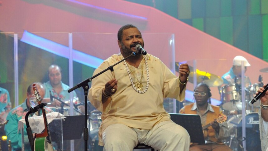 Arlindo Cruz canta sentado em um palco, vestindo roupa clara e colares, enquanto segura o microfone. Ao fundo, músicos tocam instrumentos.