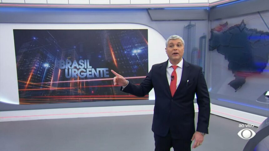 Joel Datena apresenta o Brasil Urgente em estúdio com visual urbano ao fundo e o logo do programa em destaque. A imagem representa a vitória do telejornal da Band na audiência, superando a estreia do Aqui Agora no SBT, mesmo com a presença de Geraldo Luís