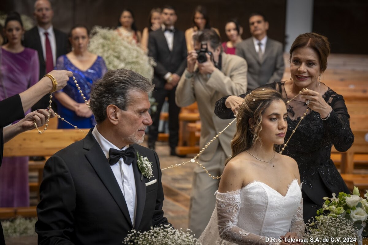 Cena do casamento de Mar em As Filhas da Senhora Garcia (SBT), mostrando a noiva e o noivo ajoelhados enquanto recebem o laço nupcial, com convidados e fotógrafo ao fundo.