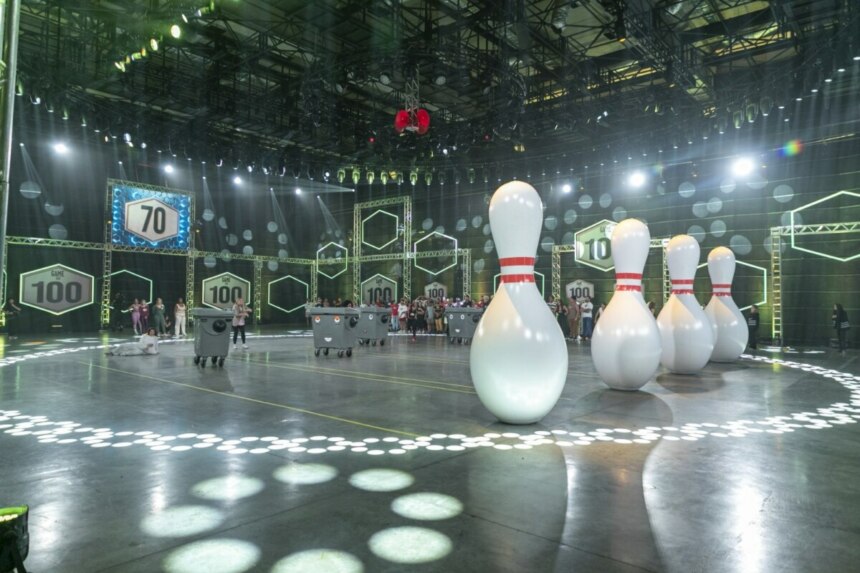 Cenário do programa Game dos 100 com 70 competidores ao fundo, grandes pinos de boliche infláveis posicionados no centro da arena e caçambas que serão usadas na prova do boliche gigante, com iluminação e painéis com o número 100 ao redor do estúdio.