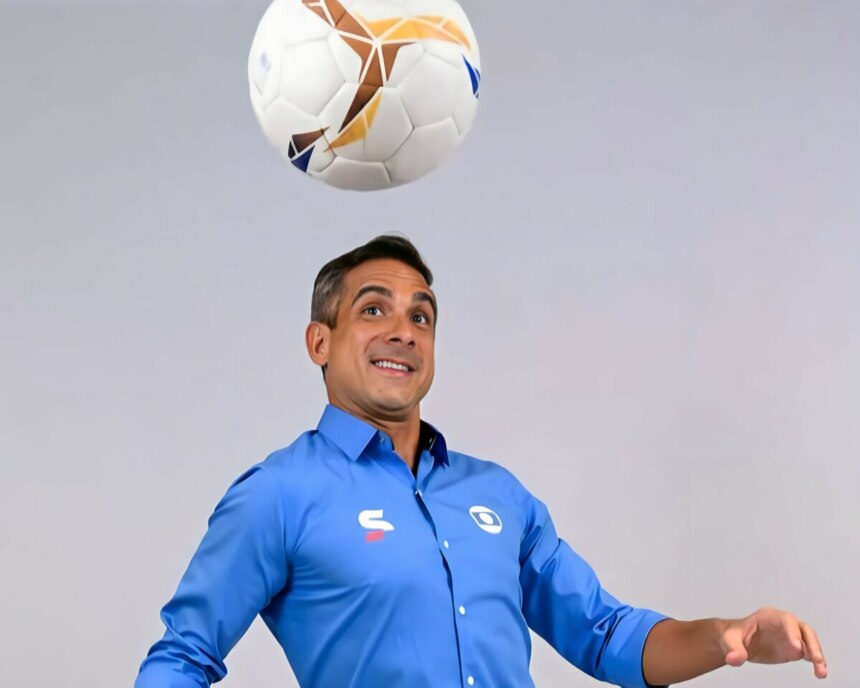 O narrador esportivo Gustavo Villani, com camisa azul, fardamento da TV Globo, olha para uma bola de futebol no ar